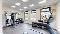Located at 970 Upper Brook Ct in Charlottesville, Virginia, this fitness center is a well-appointed, single-room exercise facility designed to support a comprehensive workout experience. The space features a drop-panel ceiling with recessed and flush-mounted lighting, providing consistent, bright illumination throughout. Large, dark-framed windows line the walls, flooding the room with natural light and offering views of the surrounding community and greenery outside.

The flooring consists of durable rubber gym matting with a speckled finish, well-suited for high-traffic fitness use. Equipment is thoughtfully arranged to maximize the available floor space. Cardio offerings include a treadmill, an elliptical trainer, and a stationary spin bike. A multi-function cable resistance machine is positioned along one mirrored wall, accompanied by an adjustable weight bench and a three-tier dumbbell rack stocked with a full range of free weights. The mirrored wall expands the sense of space and allows users to monitor their form during workouts.

Entry is through a sleek, dark-framed glass door, which connects the fitness room to an adjacent common area. The clean, neutral color palette of white walls and gray flooring gives the space a modern, professional aesthetic. Overall, the fitness center is a practical and well-equipped amenity suited for everyday strength and cardio training.