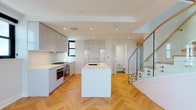 This two-floor space is defined by a cohesive modern aesthetic, with light oak herringbone parquet flooring running throughout both levels, crisp white walls, and recessed lighting that enhances the clean, contemporary atmosphere.

The main level centers around an open-plan kitchen and living area. The kitchen is well-appointed with high-gloss, handle-free cabinetry in a soft grey tone, seamless white quartz countertops, and a large waterfall island with an integrated sink. Appliances include a Bosch double oven, gas cooktop, and under-counter units. A glass-paneled staircase with a sleek railing connects the two levels, adding a striking architectural detail.

The upper floor features generously sized rooms, each continuing the herringbone flooring. Multiple rooms offer large black-framed windows with expansive views of the surrounding urban landscape. One room includes direct access to an en-suite bathroom and an adjacent walk-in closet. The bathrooms are finished with white subway tile walls, hexagonal mosaic tile floors in a warm grey tone, glass-enclosed walk-in showers with linear drains, and illuminated vanity mirrors, presenting a polished, hotel-like finish throughout.