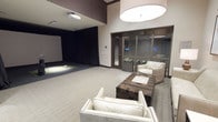 This single-floor space combines recreation and relaxation in a thoughtfully designed layout. The most distinctive feature is the indoor golf simulator, set against a large projection screen framed by dramatic black curtains, complete with a ceiling-mounted projector and a turf hitting area — ideal for year-round play and entertainment.

Adjacent to the simulator, a stylish lounge area provides a comfortable retreat. The seating arrangement includes cream-upholstered armchairs with nailhead trim detail, centered around a rich wood-and-metal coffee table adorned with a decorative accent piece. The neutral carpet and light-toned walls create a clean, contemporary backdrop throughout the space.

A double-door glass partition separates the lounge from an adjoining room, which appears to serve as a private meeting or viewing area, outfitted with its own seating and interior greenery. A large, circular pendant light anchors the lounge area, casting warm, even illumination across the space. The high two-story ceiling height in the simulator zone adds to the overall sense of openness and functionality, while maintaining a polished, upscale ambiance throughout.