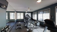 This single-floor fitness space offers a functional and inviting environment for exercise. The room is defined by its deep teal-painted walls, which contrast cleanly against white window trim and a bright white ceiling, creating a polished, focused atmosphere. Large windows line multiple walls, allowing generous amounts of natural light to fill the space and providing outdoor views that enhance the overall workout experience. The flooring features durable rubber matting, practical for both safety and equipment placement. Overhead, pendant-style light fixtures provide supplemental lighting throughout the room. The space is equipped with a variety of fitness machines, including elliptical trainers, a multi-function cable and weight machine, and a weight bench, catering to both cardiovascular and strength training needs. A mirrored wall on one side visually expands the room while allowing users to monitor their form during workouts. The layout is thoughtfully arranged to maximize usable floor space while maintaining comfortable circulation between equipment. Overall, the fitness room combines a clean aesthetic with practical amenities to support a well-rounded exercise routine.