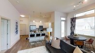 This single-story space features an open-concept layout that integrates the kitchen, dining, and living areas into one cohesive, light-filled environment. The kitchen is well-appointed with stainless steel appliances, including a French door refrigerator, microwave, and range. White shaker-style cabinetry extends to the upper walls, complemented by light stone countertops. A central island with bar seating serves as both a prep space and a casual dining area, illuminated by pendant lights that add a contemporary touch. Wood-look flooring runs throughout, providing warmth and visual continuity across the space. The living area accommodates a full-sized sofa and accent pieces, creating a comfortable and functional gathering space. A dining nook is positioned near a large window, which allows natural light to fill the area and offers a view of the outdoors. The overall aesthetic is clean and modern, characterized by a neutral color palette of whites and grays, enhanced by carefully selected furnishings and décor. The layout maximizes the use of available space, making the unit feel open and airy throughout.