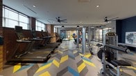 This fitness center is a thoughtfully designed, single-level facility that combines modern aesthetics with comprehensive workout functionality. One of its most striking design elements is the eye-catching chevron-patterned flooring in shades of yellow, blue, and grey, which runs throughout the entire space and lends a vibrant, energetic atmosphere.

The main workout floor is generously sized and organized into distinct zones. A dedicated cardio area lines one wall, featuring a row of treadmills, ellipticals, stair climbers, and stationary cycling bikes. Exposed brick columns punctuate this section, adding an industrial character, while horizontal windows along the upper portion of the wall allow natural light to filter in. Ceiling fans provide additional air circulation throughout.

A strength and resistance training zone is centrally located, outfitted with cable machines, multi-functional weight stations, and free weights. Industrial-style pendant lighting hangs from the ceiling, complementing the dark exposed beams overhead.

A glass-enclosed studio room with black steel-framed partition walls offers a separate space for group classes or individual training, furnished with yoga mats and weighted balls. Adjacent areas include a stretching zone with built-in bench seating and a wall-mounted television. The overall ambiance is modern, bold, and purposefully functional.