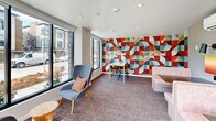 Located on 9th Ave NE in Seattle's 98115 neighborhood, this single-floor communal space combines thoughtful design with energetic artistic accents across a compact yet efficiently arranged footprint. The layout comprises a living room and an office, each distinguished by its own character while sharing a cohesive modern aesthetic.

The living room is a bright, welcoming gathering area anchored by floor-to-ceiling windows along multiple walls, flooding the space with natural light and offering views of the surrounding Seattle streetscape. Seating is varied and deliberately arranged, featuring upholstered booth-style banquettes, mid-century lounge chairs, and small side tables — accommodating both social interaction and quiet relaxation. A built-in wooden desk runs beneath a wide picture window, providing a casual workstation with neighborhood views. A striking geometric mural in bold reds, oranges, teals, and whites commands one wall, serving as a vibrant focal point. Recessed lighting and neutral carpeting ground the space.

The office features its own statement mural — an abstract, faceted design in cool blues and whites — lending a calm, creative atmosphere. Large windows continue the theme of natural light, with seating arranged around a central table for focused work or small meetings. Together, both rooms deliver a dynamic, well-lit environment suited to productivity and community engagement.