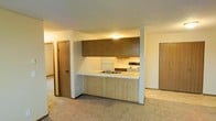 This single-floor residence is centered around an open-concept layout that seamlessly connects the kitchen and living areas. The kitchen is defined by a peninsula with a light-toned countertop and a double basin sink, offering both functional workspace and a natural divider between the cooking and living spaces. Upper and lower cabinetry feature a warm wood-grain finish with a vertical ribbed texture, adding a classic, understated character to the space. A white refrigerator and electric range are included, providing all essential appliances. Overhead, flush-mounted lighting keeps the kitchen area well-illuminated. The surrounding living space is finished with wall-to-wall carpet in a neutral tone, contributing to a comfortable, quiet atmosphere underfoot. The textured ceiling is consistent throughout, giving the space a cohesive look. Multiple interior doors are visible from the main living area, indicating access to additional rooms and storage spaces. The overall layout is practical and straightforward, with a warm, neutral color palette throughout that provides a versatile backdrop for a variety of interior styles.