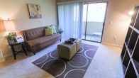 This single-floor residence offers a comfortable and well-appointed living environment designed for everyday functionality. The living area serves as the focal point of the space, featuring wall-to-wall carpeting that lends a warm and cozy feel underfoot. A large sliding glass door dominates one wall, fitted with vertical blinds that allow for adjustable natural light throughout the day. The door opens directly onto a private balcony, providing a seamless connection to the outdoors and offering additional space for relaxation. Natural light filters generously into the room, creating a bright and airy atmosphere. The layout provides ample room for comfortable furnishings, as evidenced by the generous seating arrangement and central area rug that anchors the space. Neutral wall tones complement the carpeted floors, contributing to a calm and versatile aesthetic that can accommodate a variety of décor styles. A decorative accent wall piece adds a touch of color and personality to the room. Overall, the space strikes a balance between practicality and comfort, with thoughtful details that make it a welcoming and livable environment.