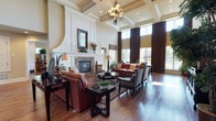 This single-story space centers around an expansive great room that immediately impresses with its soaring two-story coffered ceilings, elegantly detailed with deep box molding and cream-toned finishes. An ornate chandelier with warm-toned bulbs hangs as a focal point, complementing the refined architectural character of the room. The space is anchored by a stately stone fireplace framed by a substantial white mantel with curved architectural detailing, adding a classic and grand aesthetic. Rich, dark hardwood flooring runs throughout, providing a warm contrast to the light walls and ceiling. Floor-to-ceiling windows dressed with full-length chocolate-brown draping panels flood the room with generous natural light while offering a view of the surrounding exterior greenery. The seating area is thoughtfully arranged with leather upholstered furniture in warm cognac tones, complemented by woven accent chairs and a dark wood cocktail table with decorative accessories. The overall ambiance balances formal elegance with comfortable livability, combining traditional architectural elements with warm, inviting furnishings to create a sophisticated yet welcoming living environment.