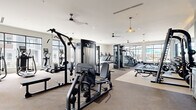 Located at 4590 Trade St in Johnstown, CO, this fitness center and yoga room is a well-appointed facility designed to support a full range of workout activities. The main fitness area is generously sized, featuring large floor-to-ceiling windows that flood the space with natural light and offer views of the surrounding community. Ceiling fans and pendant lighting complement the bright, airy atmosphere throughout.

The fitness floor is comprehensively equipped with cardio machines including treadmills, ellipticals, stair climbers, and rowing machines. A dedicated strength training zone features a multi-function cable machine, a Smith machine with bench press, a full rack of dumbbells, and additional free weight benches. A striking abstract blue-toned mural accent wall anchors the strength area, adding a stylish visual focal point. Mounted flat-screen TVs provide entertainment during workouts.

The yoga room is accessed through sleek black-framed glass sliding doors and offers a serene, open environment with light hardwood flooring, vaulted ceilings, a textured brick accent wall, and expansive windows overlooking the streetscape. Stability balls and resistance equipment are neatly stored within the room. Large glass walls between the yoga studio and the main gym maintain a sense of openness while providing a distinct, focused space for stretching and mindfulness practice.
