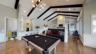 Located at 21739 Hardy Oak Blvd in San Antonio, Texas, this single-floor clubhouse delivers an impressive blend of rustic architectural character and contemporary interior design. The centerpiece of the space is the dramatic great room, where soaring cathedral ceilings are defined by bold, dark timber cross-beam trusses and geometric pendant light fixtures that cast a warm, inviting glow. Rich hardwood flooring runs throughout, tying the various areas together seamlessly. A floor-to-ceiling fireplace anchors one wall, while expansive multi-pane windows flood the room with abundant natural light and offer views of the surrounding outdoors. Distinctive Western-inspired décor, including a curated wall display of sculpted longhorn skulls in black and gold, lends the space a bold, regional character. An adjacent lounge area features vaulted ceilings with dark beams, a stone fireplace, a full-size billiards table, and a mounted flat-screen TV, creating an ideal entertainment zone. A curved quartz wet bar with bold veining, mustard-yellow cabinetry, a mosaic tile backsplash, and bar seating serves as a functional and stylish gathering point. A quieter sitting nook with wainscoting, large windows, and cowhide accents provides a more intimate retreat within the broader space.