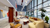 Located at 2050 Central Rd in Fort Lee, NJ, this meticulously designed residential lobby offers a generous and welcoming entry experience spanning a single expansive floor. The space is anchored by a striking concierge desk finished in veined stone, framed by rich wood paneling and a textured stone accent wall, setting a tone of understated luxury from the moment of arrival.

The main lounge area features soaring double-height ceilings adorned with a patterned geometric wallcovering in cool blue tones, complemented by a cascading pendant light fixture. Curved, neutral-toned sofas and warm cognac leather armchairs are arranged around a marble-topped coffee table, creating an inviting seating environment bathed in natural light from floor-to-ceiling curtain-wall windows that frame views of the surrounding streetscape and lush greenery.

A secondary lounge area showcases built-in dark walnut shelving flanking a sleek linear fireplace, with herringbone wood flooring adding warmth and texture. A dining area with pendant lighting and a tile-screen partition offers a refined space for residents to gather.

The hallways lead to multiple polished stainless steel elevator banks set against stone tile flooring and classic wood trim detailing. Throughout, the lobby maintains a cohesive palette of natural stone, warm wood tones, and soft neutrals, delivering a sophisticated and welcoming ambiance.