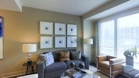 This single-floor space is thoughtfully designed to blend comfort and contemporary style. The living area serves as the centerpiece, showcasing warm, neutral-toned walls that provide an elegant backdrop for a bold grid of six framed blue agate artwork pieces, creating a striking focal point. A plush charcoal gray sofa anchors the seating arrangement, adorned with an eclectic mix of patterned and solid throw pillows in blue, brown, and geometric tones. A glass-topped coffee table adds a light, modern touch to the space. A tan leather accent chair is positioned near the window, inviting relaxation while taking in the views. Large windows dressed with sheer curtains allow generous natural light to fill the room, enhancing the open and airy ambiance. Ambient lighting is complemented by elegant floor and table lamps that add warmth during evening hours. The overall aesthetic is polished yet approachable, blending modern décor sensibilities with inviting comfort — making this a space well-suited for both everyday living and entertaining.