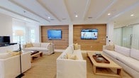 Located at 151 Du Rhu Dr in Mobile, Alabama, this meticulously designed single-floor property showcases a seamless blend of coastal warmth and modern sophistication. The expansive main living area is a standout feature, anchored by two distinctive horizontal wood-plank accent walls, each hosting wall-mounted flat-screen televisions. Plush white slipcovered sofas, natural fiber area rugs, and light wood coffee tables create an inviting, relaxed atmosphere throughout the generous seating zones. Coffered ceilings with recessed lighting add architectural depth and a sense of elevated refinement to the living spaces. A secondary sitting room offers a more intimate setting, complete with a sleek built-in fireplace flanked by large windows that allow natural light to filter in, complemented by lush indoor greenery. The open-concept kitchen and dining area are equally impressive, featuring warm wood cabinetry, marble-look surfaces, stainless steel double wall ovens, a professional-grade gas cooktop, and a large kitchen island. Pendant lantern lighting above the dining table adds a refined touch to the space. Light travertine tile flooring flows throughout the kitchen and dining areas, tying the interior palette together cohesively. Overall, the space exudes a bright, airy, and coastal-contemporary ambiance.