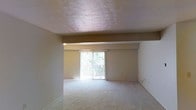 This single-floor space features an open living area finished with wall-to-wall carpeting in a neutral tone, creating a clean and understated foundation throughout. A defining feature of the room is the sliding glass door framed by vertical blinds, which fills the space with natural light and provides direct access to a private balcony overlooking lush greenery. The exterior view adds a pleasant natural backdrop to the interior. A structural beam runs horizontally across the ceiling, adding a subtle architectural detail to the room. The textured ceiling and warm, neutral-toned walls contribute to a cohesive and understated aesthetic. The layout appears open and versatile, offering flexibility in furniture arrangement. Overall, the space presents a functional and straightforward design with practical features suited for comfortable everyday living.