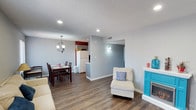 This two-floor space offers a well-organized and functional layout designed for comfortable living. The main level showcases an open-concept arrangement that seamlessly connects the living room, dining area, and kitchen. Rich hardwood-style flooring runs throughout, adding warmth and continuity to the space.

The living area features a cozy seating arrangement accented by a decorative fireplace with a distinctive turquoise door detail, lending a touch of character to the room. Recessed lighting provides consistent, even illumination throughout. An elegant chandelier hangs over the dining area, which comfortably accommodates a dark wood dining set, creating a welcoming atmosphere for meals.

The kitchen is visible from the main living space and features white cabinetry and appliances, including a full-sized refrigerator. A partial wall divides the kitchen from the dining area, maintaining an open feel while defining each zone. Neutral gray walls are complemented by crisp white trim and baseboards, creating a clean, modern aesthetic. A textured ceiling adds subtle depth to the overall design.

The second floor expands the living space further, providing additional room for rest or recreation. Throughout, the layout prioritizes both practicality and an inviting ambiance suited for daily living.