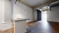Located on East Broad Street in Richmond, Virginia, this single-floor loft offers a compelling blend of industrial character and modern functionality. The open layout is defined by rich, stained concrete floors that extend throughout, lending warmth and texture to the space. Exposed concrete ceilings with visible structural elements, ductwork, and conduit reinforce the raw, loft-style aesthetic, while ceiling fans provide practical airflow across the open floor plan.

The main living and kitchen area serves as the heart of the space. A peninsula-style kitchen counter with a granite surface and built-in sink anchors the kitchen zone, complemented by stainless steel appliances, a wall oven, and light wood cabinetry. Pendant lighting above the counter adds a refined touch. Painted brick columns punctuate the open area, adding architectural interest and a nod to the building's historic character. Natural light filters in from the far end of the space, brightening the hallway that connects the primary areas.

The bathroom continues the industrial theme with polished concrete flooring, a floating wood vanity with a vessel sink, and clean white walls. Chrome fixtures and a towel bar complete the utilitarian yet stylish finish. Overall, this space offers a cohesive industrial-modern environment well-suited for a variety of uses.