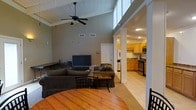 This single-story space is defined by its striking vaulted ceiling in the main living area, which is finished with white-painted wood planks and fitted with a ceiling fan, creating an airy, open atmosphere. The tall ceiling height adds a sense of spaciousness well beyond what the footprint might suggest. The living room is carpeted and comfortably furnished with dark upholstered sofas and a coffee table, centered around a flat-screen television. A wooden dining table with chairs occupies the foreground, serving as a natural transition between the living and dining areas. Decorative columns frame the passage into the kitchen, adding a subtle architectural detail. The kitchen features warm wood cabinetry, tile flooring, and is equipped with stainless steel appliances including a range and microwave. A hallway visible from the living room suggests additional rooms beyond the main open-plan area. The overall ambiance is casual and functional, with a neutral gray color palette throughout the living space contrasting with the warmer tones of the kitchen cabinetry and dining table.