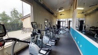 This fitness center is a thoughtfully designed, single-floor facility built to support a comprehensive workout experience. The main exercise area is long and open, featuring high vaulted ceilings that create a spacious, breathable atmosphere. Multiple ceiling fans run the length of the room, ensuring comfortable airflow during workouts.

Natural light pours in through large windows along the main wall, offering outdoor views of a patio area with seating and surrounding greenery — a welcoming visual backdrop during exercise. The flooring transitions between durable rubber matting and carpeted zones, providing appropriate surfaces for different types of training.

A vibrant teal accent wall runs along one side of the main room, adding a bold pop of color to the otherwise neutral beige and gray palette. Full-length mirrors line the opposite wall, enhancing the sense of space and allowing users to monitor their form.

The cardio section is well-stocked with a row of elliptical trainers and treadmills, each equipped with digital display screens. A multi-station cable strength machine is positioned toward the rear of the space, alongside a freestanding dumbbell rack with a full range of weights. A separate, carpeted room with mirrored walls provides a dedicated area for stretching or additional training. Exposed ductwork along the ceiling adds an industrial design element throughout.