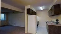 This single-floor unit offers a straightforward and functional layout with an open connection between the kitchen and living areas. The kitchen is equipped with essential appliances, including a full-size white refrigerator, an electric range, and additional counter space along the perimeter. Dark wood upper cabinets are mounted above the refrigerator on a structural column, providing storage while defining the boundary between the kitchen and the adjoining living space. The kitchen features tile or vinyl flooring, which transitions into carpeting in the open living area. Walls are finished in a neutral off-white tone, contributing to a clean and simple aesthetic throughout. Overhead fluorescent lighting illuminates the kitchen area, ensuring practical visibility. The textured ceiling adds a subtle architectural detail consistent with the overall utilitarian design of the space. The living area, visible through the open passthrough, offers a generous footprint with carpeted flooring and a neutral color palette. Overall, the space presents a no-frills, functional environment well-suited for everyday living.