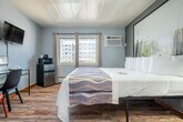 Located at 3177 E Washington Ave in Madison, Wisconsin, this single-floor unit offers a thoughtfully designed layout that maximizes functionality within a compact footprint. The main bedroom area is generously sized relative to the overall space, featuring warm wood-style flooring, cool gray walls, and a paneled white ceiling that keeps the room feeling bright and clean. Natural light enters through windows fitted with curtains, and a wall-mounted air conditioning unit ensures year-round comfort. The room is furnished with a king-sized bed backed by a large nature-inspired wall art piece, a sleek glass-top nightstand with a modern lamp, and a wall-mounted flat-screen television. A minimalist desk with seating provides a functional workspace, while a compact refrigerator and microwave add convenient in-room amenities. A full-length mirror and clothing rack offer practical storage solutions. The bathroom features classic mosaic tile flooring in soft earth tones, a combined shower and tub with a curtain, a single vanity with a wide countertop, and ample towel storage. The overall aesthetic is modern and understated, blending simple furnishings with clean lines for a comfortable and practical living experience.