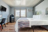 Located at 3177 E Washington Ave in Madison, Wisconsin, this single-floor unit offers a thoughtfully designed layout that maximizes functionality within a compact footprint. The main bedroom area is generously sized relative to the overall space, featuring warm wood-style flooring, cool gray walls, and a paneled white ceiling that keeps the room feeling bright and clean. Natural light enters through windows fitted with curtains, and a wall-mounted air conditioning unit ensures year-round comfort. The room is furnished with a king-sized bed backed by a large nature-inspired wall art piece, a sleek glass-top nightstand with a modern lamp, and a wall-mounted flat-screen television. A minimalist desk with seating provides a functional workspace, while a compact refrigerator and microwave add convenient in-room amenities. A full-length mirror and clothing rack offer practical storage solutions. The bathroom features classic mosaic tile flooring in soft earth tones, a combined shower and tub with a curtain, a single vanity with a wide countertop, and ample towel storage. The overall aesthetic is modern and understated, blending simple furnishings with clean lines for a comfortable and practical living experience.