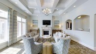 This beautifully designed single-floor space exudes timeless elegance and refined comfort. The centerpiece of the living area is a classic white-mantled fireplace, flanked by warm wood sideboards and framed by a pair of serene, coastal-inspired landscape paintings. A flat-screen television is mounted above the mantle, seamlessly blending modern functionality with traditional design. The ceiling commands immediate attention with its intricate coffered detailing, finished in soft white and accented by recessed lighting and a statement chandelier that casts a warm, inviting glow throughout the room. The seating arrangement is thoughtfully curated, featuring neutral-toned sofas paired with patterned accent chairs in a soft blue and gold geometric motif, centered around a delicate glass and metal coffee table and a gold side table. Large windows along the perimeter invite an abundance of natural light, enhancing the airy, open ambiance of the space. An arched architectural niche adds a graceful structural detail to one wall, lending depth and character. The overall palette of soft whites, warm golds, and muted blues creates a cohesive, sophisticated atmosphere that is both welcoming and visually refined.