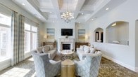 This beautifully designed single-floor space exudes timeless elegance and refined comfort. The centerpiece of the living area is a classic white-mantled fireplace, flanked by warm wood sideboards and framed by a pair of serene, coastal-inspired landscape paintings. A flat-screen television is mounted above the mantle, seamlessly blending modern functionality with traditional design. The ceiling commands immediate attention with its intricate coffered detailing, finished in soft white and accented by recessed lighting and a statement chandelier that casts a warm, inviting glow throughout the room. The seating arrangement is thoughtfully curated, featuring neutral-toned sofas paired with patterned accent chairs in a soft blue and gold geometric motif, centered around a delicate glass and metal coffee table and a gold side table. Large windows along the perimeter invite an abundance of natural light, enhancing the airy, open ambiance of the space. An arched architectural niche adds a graceful structural detail to one wall, lending depth and character. The overall palette of soft whites, warm golds, and muted blues creates a cohesive, sophisticated atmosphere that is both welcoming and visually refined.