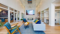 Located at 920 Yorkmont Ridge Lane in Fort Mill, South Carolina, this single-story clubhouse delivers a polished, contemporary design defined by dramatic double-height ceilings and an abundance of natural light streaming through expansive floor-to-ceiling glass walls and doors. Light hardwood flooring runs throughout, creating a cohesive and airy aesthetic that connects each area of the space.

The main lounge serves as the centerpiece, anchored by a sleek, tile-clad linear fireplace topped by a large mounted display screen. Comfortable seating arrangements, including mid-century-inspired teal accent chairs and neutral sofas accented with bold blue and yellow throw pillows, are arranged around patterned area rugs, fostering an inviting atmosphere for gathering or relaxing.

Adjacent to the main lounge, a secondary seating area provides additional casual lounging space, with glass-partitioned walls offering views into neighboring rooms while maintaining an open feel. A glass-enclosed conference room features a statement chandelier and a full boardroom table, ideal for professional meetings.

A kitchenette area includes a central island with bar seating, stainless steel appliances, and a decorative patterned accent wall, adding a playful design element. Throughout the space, generous glazing frames outdoor greenery, connecting the interior to the natural surroundings and enhancing the bright, welcoming ambiance.