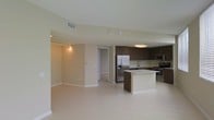 This single-floor space showcases a thoughtfully designed open-concept layout that integrates the kitchen and living areas into one cohesive environment. The kitchen is a standout feature, outfitted with rich dark brown cabinetry that extends to the upper walls, providing ample storage. Stainless steel appliances, including a French door refrigerator with an ice and water dispenser, a built-in microwave, and a range with oven, lend a polished, contemporary feel. A central kitchen island with a light-toned countertop serves as both a prep surface and a natural dividing point between the kitchen and the open living space. The sink, positioned along the back counter, overlooks the kitchen with practical accessibility. Light-toned tile flooring extends throughout the space, enhancing the sense of openness and reflecting the overhead lighting effectively. The living area is generously proportioned, with neutral beige walls and white baseboards contributing to a clean, understated aesthetic. Track lighting in the living zone offers directional illumination, while a flush-mount ceiling fixture brightens the kitchen. A doorway visible from the living area suggests access to additional rooms within the unit.