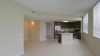 This single-floor space showcases a thoughtfully designed open-concept layout that integrates the kitchen and living areas into one cohesive environment. The kitchen is a standout feature, outfitted with rich dark brown cabinetry that extends to the upper walls, providing ample storage. Stainless steel appliances, including a French door refrigerator with an ice and water dispenser, a built-in microwave, and a range with oven, lend a polished, contemporary feel. A central kitchen island with a light-toned countertop serves as both a prep surface and a natural dividing point between the kitchen and the open living space. The sink, positioned along the back counter, overlooks the kitchen with practical accessibility. Light-toned tile flooring extends throughout the space, enhancing the sense of openness and reflecting the overhead lighting effectively. The living area is generously proportioned, with neutral beige walls and white baseboards contributing to a clean, understated aesthetic. Track lighting in the living zone offers directional illumination, while a flush-mount ceiling fixture brightens the kitchen. A doorway visible from the living area suggests access to additional rooms within the unit.