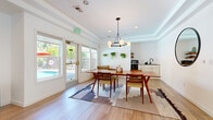 This thoughtfully designed single-floor amenity space blends mid-century modern aesthetics with contemporary functionality across several well-appointed areas. The main gathering space centers around a warm-toned dining area highlighted by a striking globe chandelier suspended beneath a tray ceiling, complemented by light hardwood floors that flow throughout. A built-in kitchenette with white cabinetry, a dark countertop, and open shelving adorned with hanging greenery provides convenient refreshment access, while French doors open directly to an outdoor pool area, allowing natural light to pour in. A white brick accent wall adds texture and character to the dining space. Adjacent to the dining area, a comfortable lounge features a caramel leather sectional sofa, a wall-mounted flat-screen television, and additional windows that frame lush outdoor views. The welcoming entry corridor connects all areas seamlessly, offering a bright, airy transition with glass-paneled doors and clean white walls. A dedicated fitness room, finished with teal accent walls and rubber flooring, is fully equipped with a multi-station cable machine, treadmill, stationary bike, adjustable bench, and free weights. A private office with teal walls and glass partitioning rounds out the space, offering a focused work environment within this versatile and well-curated facility.