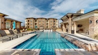 This residential community showcases an impressive array of outdoor amenities designed for comfortable, resort-style living. At the heart of the exterior is a generously sized swimming pool with crystal-clear water, surrounded by a travertine-style pool deck lined with lounge chairs and shaded tiki-style umbrellas — ideal for relaxing or socializing outdoors. An adjacent covered cabana area, constructed with matching brick columns and a pitched roof, provides a shaded retreat overlooking the pool.

The surrounding courtyard features broad, multi-toned paver walkways that connect the various amenity areas, creating a cohesive and polished outdoor environment. Landscaped beds with ornamental grasses and seasonal plantings add texture and color throughout the grounds, while a manicured lawn area provides open green space suitable for outdoor games and activities.

A pergola-covered entry corridor leads residents through attractively landscaped garden beds framed by decorative iron fencing, offering a welcoming and well-maintained pathway between buildings. The multi-story brick residential buildings rise prominently in the background, featuring balconies and large windows that overlook the amenity areas below. The overall architectural style is classic and traditional, with warm brick exteriors, dark hip roofs, and refined detailing that lend a sense of quality and permanence to the community.
