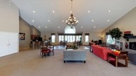 This single-floor communal space is defined by its impressive vaulted ceiling, which creates an open, airy atmosphere throughout. A wrought-iron chandelier with candle-style lights serves as a central focal point, complemented by recessed lighting that ensures consistent illumination across the entire area. A large arched window flanked by additional windows along the far wall floods the interior with natural light, enhancing the welcoming ambiance.

The layout is thoughtfully divided into functional zones. A comfortable lounge area anchors the center of the space, furnished with a light gray sofa and a bold red sectional arranged around a low coffee table. Adjacent to this is a casual dining area featuring dark wood tables and red chairs, providing ample seating for gatherings.

Toward the rear left, a kitchenette with dark cabinetry, a counter with bar-height seating, and visible appliances offers convenient refreshment access. The flooring transitions from hard surface near the dining and kitchen zones to carpet in the lounge area, subtly delineating each space while maintaining a cohesive overall design.