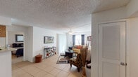 This single-floor residence offers a thoughtfully arranged layout that balances comfort and functionality across its key living areas. The living room serves as the heart of the home, featuring light tile flooring, a ceiling fan, and large windows dressed with dark curtains that allow ample natural light to fill the space. A curved sectional sofa, glass-topped coffee table, and decorative accents create a welcoming and stylish atmosphere.

Adjacent to the living area, a compact dining nook features a glass-top round table with dark wood chairs, set against a warm-toned accent wall with decorative wall art, adding a distinctive visual focal point near the entry door.

The galley-style kitchen is efficiently designed with warm oak cabinetry lining both sides, offering generous storage space. It is equipped with a double sink, a coil-top range, a refrigerator, and a dishwasher, making it a fully functional cooking space.

The bedrooms provide comfortable retreats, with one featuring hardwood-style flooring, a natural light window, and space for a work desk, while another offers carpeted flooring and a decorative wall mirror. The bathroom includes a vanity with a framed mirror, warm lighting, and a tiled shower-tub combination, completing this well-rounded residence.