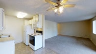 This single-floor residence features an open-concept layout that seamlessly connects the kitchen and living area, creating a practical and cohesive living environment. The kitchen is equipped with white cabinetry, a full-size refrigerator, an electric range with oven, and a built-in microwave, all arranged efficiently along a defining half-wall that subtly separates the cooking space from the main living area. The tile flooring in the kitchen provides a durable, easy-to-clean surface, while the adjacent living area transitions to wall-to-wall carpeting, adding warmth and comfort underfoot. A ceiling fan with an integrated light fixture is centrally positioned in the living space, offering both air circulation and ambient lighting. The textured ceilings and neutral wall tones throughout contribute to a clean, understated aesthetic. The open sightlines between the kitchen and living room enhance the sense of space, making the layout feel airy and functional. Overall, the unit presents a straightforward, well-organized floor plan suited for efficient everyday living.