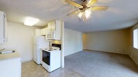 This single-floor residence features an open-concept layout that seamlessly connects the kitchen and living area, creating a practical and cohesive living environment. The kitchen is equipped with white cabinetry, a full-size refrigerator, an electric range with oven, and a built-in microwave, all arranged efficiently along a defining half-wall that subtly separates the cooking space from the main living area. The tile flooring in the kitchen provides a durable, easy-to-clean surface, while the adjacent living area transitions to wall-to-wall carpeting, adding warmth and comfort underfoot. A ceiling fan with an integrated light fixture is centrally positioned in the living space, offering both air circulation and ambient lighting. The textured ceilings and neutral wall tones throughout contribute to a clean, understated aesthetic. The open sightlines between the kitchen and living room enhance the sense of space, making the layout feel airy and functional. Overall, the unit presents a straightforward, well-organized floor plan suited for efficient everyday living.