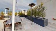 This contemporary rooftop terrace offers an impressive outdoor retreat designed for both relaxation and social gatherings. The expansive space is finished with large-format porcelain tile pavers throughout, lending a clean, modern aesthetic underfoot. A standout feature is the warm-toned wood built-in bench and platform seating area, which serves as a central gathering point and frames sweeping panoramic views of the city skyline beyond the parapet walls. Overhead, strands of bistro string lights crisscross the terrace, creating an inviting ambiance after dark. A dedicated outdoor dining area is outfitted with a long communal table and seating, accompanied by multiple freestanding patio heaters that extend the usability of the space across all seasons. Thoughtfully positioned raised planters in a matte dark finish are filled with slender ornamental trees, introducing greenery and a sense of privacy. A sleek access door provides seamless entry to and from the building's interior. The overall design balances minimalist architecture with carefully curated furnishings and plantings, resulting in a refined urban outdoor space that capitalizes on its elevated vantage point and open sky exposure.
