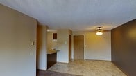 This single-story space features an open-concept layout that seamlessly connects the living area to the kitchen. The living room is finished with carpet and transitions into tile flooring toward the entryway and kitchen zones. A ceiling fan with a light fixture is centrally positioned in the living area, providing both ambient lighting and air circulation. The textured popcorn ceiling runs throughout the space, adding a classic residential character.

The kitchen is partially separated by a raised breakfast bar or counter, offering a casual dining or workspace surface while maintaining visual openness to the living area. Dark-toned cabinetry is visible in the kitchen, complemented by an overhead cabinet, providing functional storage. A door near the kitchen area likely leads to an additional room or closet, while a solid wood-toned entry door anchors the main entrance of the space.

The neutral, warm beige wall tones create a cohesive look throughout, and the layout is practical and straightforward. Light switch plates are visible near the entry, and the overall design reflects a functional, no-frills interior suited for everyday living.