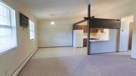 This single-floor space presents an open-concept layout that seamlessly connects the living, dining, and kitchen areas into one cohesive environment. The living and dining area is generously sized, featuring neutral-toned carpet underfoot that adds warmth and comfort to the space. Soft, cream-colored walls keep the interior feeling bright and airy, complemented by a window that allows natural light to filter in. A pendant light fixture hangs overhead, adding a simple focal point to the dining area.

The kitchen is defined by a half-wall breakfast bar with a light-colored countertop, supported by dark structural posts that create a visual boundary between the kitchen and the main living area. Dark wood cabinetry lines the upper and lower kitchen walls, providing ample storage, while a white refrigerator and under-cabinet range hood complete the functional setup. The kitchen features hard flooring that transitions cleanly into the carpeted living space.

A baseboard heater runs along the lower wall of the living area, indicating practical heating infrastructure. The overall ambiance of the space is clean, straightforward, and functional, with a neutral palette throughout that provides a versatile backdrop suitable for a variety of furnishing styles.