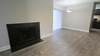 This single-floor space offers a clean, modern aesthetic defined by warm wood-style flooring that flows seamlessly throughout the open layout. A striking black-framed fireplace serves as a focal point in the main living area, providing both a functional heating element and a bold design statement against the neutral, light-gray walls. The space benefits from an abundance of natural light streaming through a large window dressed with vertical blinds, creating a bright and airy atmosphere. Adjacent to the main living area, a dedicated dining zone is highlighted by a stylish geometric pendant light fixture, adding a contemporary touch to the overall design. Crisp white baseboards frame the room, complementing the understated color palette and contributing to a polished, well-finished appearance. The open-concept arrangement between the living and dining areas promotes a sense of spaciousness and ease of movement, making the layout both practical and inviting. Overall, the space presents a well-maintained, move-in-ready environment with a cohesive modern design that balances comfort with clean architectural details.