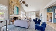 Located at 4369 S Quebec St in Denver, Colorado, this single-story clubhouse is designed to impress with its dramatic architectural features and thoughtfully curated interiors. The centerpiece of the main lounge is a floor-to-ceiling stone-clad fireplace, extending the full height of the steeply vaulted ceiling and anchored by a bold teal artwork that draws the eye upward. Geometric pendant chandeliers in a dark metal finish hang overhead, adding a contemporary industrial accent against the bright white ceiling.

The expansive lounge area is furnished with a mix of soft lavender sectional sofas, neutral accent chairs, and rich cobalt blue velvet armchairs, creating a layered, inviting palette. A patterned area rug with circular motifs grounds the central seating arrangement, which is completed by glass and metal coffee tables. Multiple French doors and transom windows line the perimeter, flooding the space with natural light and offering views of the surrounding greenery.

A secondary lounge area transitions seamlessly from the main space, featuring high-top bar tables with modern white stools positioned beneath a gallery wall of abstract artwork. An adjacent office area with workstations and additional French doors is visible through a wide architectural opening, reinforcing the open, connected layout throughout.