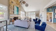 Located at 4369 S Quebec St in Denver, Colorado, this single-story clubhouse is designed to impress with its dramatic architectural features and thoughtfully curated interiors. The centerpiece of the main lounge is a floor-to-ceiling stone-clad fireplace, extending the full height of the steeply vaulted ceiling and anchored by a bold teal artwork that draws the eye upward. Geometric pendant chandeliers in a dark metal finish hang overhead, adding a contemporary industrial accent against the bright white ceiling.

The expansive lounge area is furnished with a mix of soft lavender sectional sofas, neutral accent chairs, and rich cobalt blue velvet armchairs, creating a layered, inviting palette. A patterned area rug with circular motifs grounds the central seating arrangement, which is completed by glass and metal coffee tables. Multiple French doors and transom windows line the perimeter, flooding the space with natural light and offering views of the surrounding greenery.

A secondary lounge area transitions seamlessly from the main space, featuring high-top bar tables with modern white stools positioned beneath a gallery wall of abstract artwork. An adjacent office area with workstations and additional French doors is visible through a wide architectural opening, reinforcing the open, connected layout throughout.