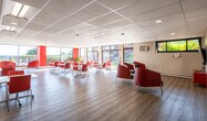 This single-floor facility is designed as a welcoming, multi-functional communal space with a clean, modern aesthetic. The interior is unified by a bold red-and-white color scheme, reflected in the furnishings and architectural accents throughout. Light hardwood-style flooring runs continuously across the open areas, complementing the white drop-panel ceilings fitted with recessed and linear lighting fixtures.

The main gathering area is generously proportioned, featuring multiple white rectangular tables on casters paired with red stacking chairs, allowing for flexible arrangement to accommodate various group sizes and activities. Expansive floor-to-ceiling windows and glass doors line the perimeter, inviting abundant natural light and offering clear views of the surrounding greenery and outdoor grounds.

A lounge area provides a more relaxed setting, furnished with bold red and black upholstered armchairs arranged for conversation. A separate, more intimate room serves as a media or recreation space, equipped with a wall-mounted flat-screen television, additional red lounge chairs, and a small storage console.

A reception or service counter area connects functional zones within the space, complete with pass-through windows and access to additional rooms. Safety features including fire extinguishers and clearly marked exits are present throughout. Overall, the space projects an energetic, organized, and community-oriented atmosphere suited for gatherings, programming, and everyday use.