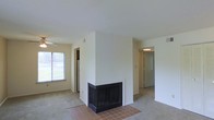 This single-floor space is designed with an open and functional layout, centered around a standout architectural feature: a freestanding, two-sided fireplace with a dark surround and tiled hearth in warm terracotta tones. The fireplace serves as a natural room divider, adding both character and practicality to the living area.

The floors transition between tile at the hearth and neutral carpeting throughout the remaining living spaces, lending a cozy, understated feel. The walls are finished in clean white, contributing to a bright and airy atmosphere. A window along one wall allows natural light to filter into the space, offering a view of greenery outside.

The layout connects the main living area to additional rooms via open doorways and corridors, suggesting a well-organized flow between spaces. A glimpse of what appears to be a kitchen or utility area is visible in the background, alongside a hallway leading to further rooms.

The ceilings are smooth and standard height, and the overall aesthetic is simple and neutral, providing a blank canvas well-suited for a variety of interior styles and furnishing preferences.