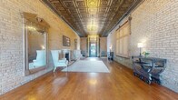 Located at 330 E Ninth St in Saint Paul, Minnesota, this historic single-floor space offers a generously sized, open layout rich in architectural character. The interior is immediately distinguished by its stunning ornate pressed-tin ceiling, adorned with intricate geometric and floral patterns and illuminated by elegant crystal chandeliers that cast warm light throughout the room. Exposed brick walls in a classic pale yellow tone run the length of the space, lending a timeless industrial-historic aesthetic that is both refined and rustic. Wide-plank hardwood floors in rich warm tones anchor the interior, adding continuity and warmth. A standout architectural feature is the decorative marble fireplace with an ornate cast-iron surround, creating a natural focal point in the main room. Large multi-pane entry doors allow abundant natural light to flood the interior, while classic cast-iron radiators speak to the building's historic origins. A wooden staircase with dark metal railings connects to an additional level, accompanied by a reception desk area that suggests strong utility as a venue or professional space. Oversized sliding wooden barn-style panels and exposed structural beams in the rear section add to the layered character of the space. The overall ambiance blends 19th-century historic grandeur with versatile, event-ready functionality.