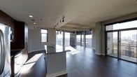 This contemporary single-floor space showcases a thoughtfully designed open-concept layout that seamlessly integrates the kitchen and living areas into one cohesive environment. Dark hardwood flooring runs throughout, providing a rich, grounding contrast to the bright, airy atmosphere created by the expansive floor-to-ceiling windows that line the exterior walls. These oversized windows flood the interior with natural light while framing sweeping panoramic views of the surrounding cityscape, making the space feel open and connected to the urban landscape beyond.

The kitchen is anchored by a central island featuring granite countertops with subtle veining, offering both additional workspace and casual seating. Modern pendant lighting and a sleek track lighting system overhead provide functional illumination, complementing the clean, minimalist aesthetic. The cabinetry is finished in a contemporary style, contributing to the overall streamlined look.

The living area benefits greatly from the corner window configuration, maximizing light exposure and views from multiple angles. Smooth white ceilings, clean architectural columns, and crisp white baseboards complete the refined, modern interior. The overall ambiance is polished and sophisticated, ideal for those who appreciate contemporary design paired with an impressive urban outlook.