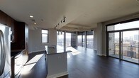 This contemporary single-floor space showcases a thoughtfully designed open-concept layout that seamlessly integrates the kitchen and living areas into one cohesive environment. Dark hardwood flooring runs throughout, providing a rich, grounding contrast to the bright, airy atmosphere created by the expansive floor-to-ceiling windows that line the exterior walls. These oversized windows flood the interior with natural light while framing sweeping panoramic views of the surrounding cityscape, making the space feel open and connected to the urban landscape beyond.

The kitchen is anchored by a central island featuring granite countertops with subtle veining, offering both additional workspace and casual seating. Modern pendant lighting and a sleek track lighting system overhead provide functional illumination, complementing the clean, minimalist aesthetic. The cabinetry is finished in a contemporary style, contributing to the overall streamlined look.

The living area benefits greatly from the corner window configuration, maximizing light exposure and views from multiple angles. Smooth white ceilings, clean architectural columns, and crisp white baseboards complete the refined, modern interior. The overall ambiance is polished and sophisticated, ideal for those who appreciate contemporary design paired with an impressive urban outlook.