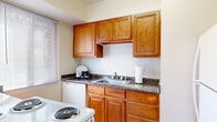 This single-floor unit offers a practical and well-organized layout connecting several distinct living spaces. The entry foyer features a dark wood accent floor that transitions into wall-to-wall carpeting extending throughout the living areas, hallway, and bedrooms. The open living area is generously proportioned, featuring neutral walls with a contrasting blue accent wall, a large window with vertical blinds that allows ample natural light, and ceiling ventilation throughout.

The kitchen is a separate, dedicated space finished with warm honey-toned raised-panel cabinetry that reaches toward the ceiling, providing substantial storage. Dark granite-style countertops offer contrast against the white appliances, which include a coil-burner range, dishwasher, and full-size refrigerator. Light wood-look flooring gives the kitchen a clean, bright feel distinct from the rest of the unit.

The bedrooms are carpeted and well-lit by individual windows with horizontal blinds. Each room features clean white walls and trim, offering a neutral backdrop adaptable to various uses. The bathroom is finished with warm beige tile walls and flooring, a modern floating-style vanity with a rectangular basin sink, a mirrored medicine cabinet, and a windowed shower-tub combination, giving the space a clean and updated appearance.