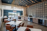 Located at 240 Avenida Vista Montana in San Clemente, California, this single-level community clubhouse draws inspiration from the area's coastal character and delivers a vibrant, well-appointed gathering space. The interior makes an immediate impression with its signature ceiling treatment—thick nautical ropes draped dramatically overhead, paired with Edison bulb pendants that cast warm, ambient light throughout. A bold teal accent ceiling anchors the central zone and complements the deep blue feature wall at the far end of the space.

The open-plan layout flows seamlessly between a casual dining area, a relaxed lounge, and a kitchenette. The dining zone features multiple white-topped tables with tan leather chairs, surrounded by large windows that flood the room with natural light and frame views of the landscaped exterior. Curated artwork, including oversized photography and neon-inspired pieces, adds visual energy to the walls.

The kitchenette is outfitted with modern appliances, light wood cabinetry, and a striking black-and-white patterned tile accent wall. The lounge area offers plush seating and a large flat-screen TV, ideal for casual gatherings. Polished concrete floors run throughout, tying the spaces together with a clean, contemporary finish. Expansive sliding glass doors open directly to the outdoor pool terrace, blurring the line between indoor comfort and Southern California sunshine.