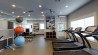 This fitness center offers a thoughtfully organized workout environment designed to accommodate a range of exercise routines. The space is outfitted with multiple SportsArt treadmills along one side, providing dedicated cardio stations for users. On the opposite end, a dumbbell rack and various free weights are arranged alongside large exercise balls in multiple sizes, supporting strength and flexibility training.

The room features neutral gray walls adorned with abstract artwork, lending a clean and modern aesthetic. Recessed lighting is distributed throughout the ceiling, supplemented by multiple ceiling fans that help maintain airflow and comfort during workouts. A wall-mounted mini-split air conditioning unit provides additional climate control. Natural light enters through several windows fitted with blinds, keeping the atmosphere bright without glare.

A wooden storage unit near the center of the room offers convenient organization for smaller equipment and accessories. A clearly marked exit door at the rear provides easy access. Overall, the space balances functionality with a polished, welcoming design suitable for everyday fitness use.