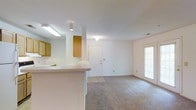 This single-floor residence offers a clean, functional layout designed for comfortable everyday living. The main living area is generously proportioned, featuring neutral wall-to-wall carpeting and crisp white walls that create a bright, airy atmosphere. A door with sidelight windows allows natural light to filter into the living space, while blinds provide privacy when needed. A hallway extends from the main living area, connecting to additional rooms at the rear of the unit, with overhead lighting guiding the way.

The kitchen is positioned adjacent to the living area and features a peninsula-style breakfast bar that serves as a natural divider between the two spaces. Warm honey-toned wood cabinetry lines the kitchen walls, complemented by a light-colored countertop. Overhead fluorescent lighting ensures the kitchen is well-illuminated for meal preparation. A front entry door is visible from the kitchen area, offering a convenient and welcoming point of entry.

Throughout the unit, the textured white ceilings and consistent neutral carpeting tie each area together cohesively. The overall design is straightforward and practical, offering an easily customizable canvas well-suited for a variety of living arrangements.