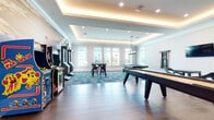 Located at 1703 Pope Rd in Bradenton, Florida, this single-floor entertainment and game room is designed as a dynamic recreational space with a modern, polished aesthetic. The room is defined by its tray ceilings with warm cove lighting that complements recessed fixtures, creating a layered, inviting ambiance throughout. Wide-plank hardwood-style flooring covers much of the space, transitioning to vibrant teal and gray patterned carpet tiles in designated seating and activity zones, adding visual interest and defining distinct areas within the open layout.

The space is comprehensively equipped for recreation, featuring multiple shuffleboard tables, a pool table, and a collection of classic arcade machines — including Pac-Man and a dart game cabinet — lined along a dramatic stadium-themed wall mural that serves as a bold focal point. Wall-mounted flat-screen televisions are positioned for easy viewing from the seating areas, which include high-top tables with modern bar-height chairs.

Large windows along the perimeter flood the room with natural light and offer views of the surrounding outdoor greenery. A pendant chandelier adds a refined touch above the central gathering area. The overall design balances entertainment functionality with contemporary style, making it a well-appointed communal amenity space.